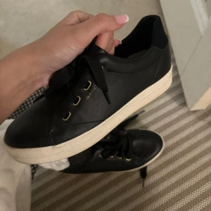 Svarta sneakers från Gant - Snygga svarta sneakers från Gant i skinn med vita sulor. De har en klassisk design med snörning och guldiga detaljer vid snörhålen. Perfekta för både vardag och festliga tillfällen. Skorna är i bra skick och redo för nya äventyr!