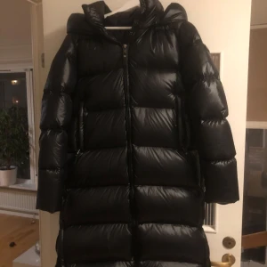 Svart dunjacka från Cedrico - Säljer en snygg och varm svart dunjacka från Cedrico i storlek S. Jackan har en lång modell med huva och dragkedja framtill. Perfekt för kalla vinterdagar när du vill hålla dig varm och stilren. 