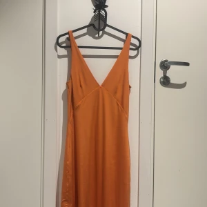 Orange axelbandslös långklänning - Säljer en snygg orange långklänning med axelband och djup v-ringning. Perfekt för sommarens fester eller en dag på stranden eller som balklänning. Klänningen har en härlig flowy känsla och är gjord i ett lätt material som känns skönt mot huden.