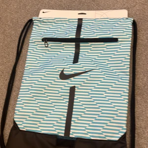 Blåvit randig gympåse från Nike - Säljer en snygg blåvit randig gympåse från Nike. Den har ett coolt sicksackmönster och en praktisk dragkedja framtill. Perfekt för träning eller som en lätt ryggsäck till vardags. Den är i nytt skick och redo för användning!