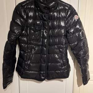 Säljer en snygg svart dunjacka från Moncler i mycket bra skick. Jackan har en glansig finish och är perfekt för kalla vinterdagar. Den har en dragkedja framtill och en praktisk bröstficka med knapp. Moncler-loggan syns på ärmen. Perfekt för dig som vill hålla stilen i vinter! ❄️