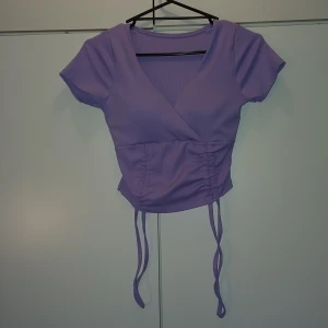 Kort lila topp - En kort lila topp som passar till alla tillfällen. Den har använts en gång och har därför inte prislappen kvar. Den går att justera i längd. Det har fastsydda ilägg i. Den köptes på en marknad därmed finns inget märke på den.