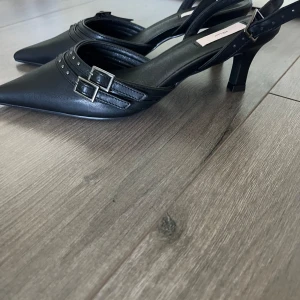 Svarta pumps med spetsig tå från Nelly - Snygga svarta pumps från Nelly med spetsig tå och stiliga spännen. De har en elegant design med dubbla remmar och små nitar som ger en extra touch. Aldrig använt pga av fel storlek. Orginalpris ca 500 Perfekta för fest eller en utekväll!