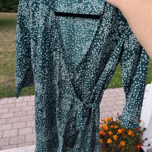 Mörkgrön omlott-klänning - En mörkgrön knälång klänning med små vita blad från Vero Moda. Klänningen läggs omlott och knyts i midjan på vänster sida. Den har volangkant längst ned samt trekvartsärm 🌟 Sann till storleken 💞  (ALDRIG ANVÄND)  Längd: 95 cm Axel: 36 cm