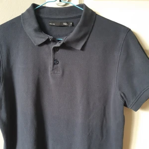 Svart pikétröja från 15% - Säljer en stilren marinblå pikétröja från lager 157. Den har en klassisk krage och knappar framtill. Perfekt för både vardag och lite mer uppklädda tillfällen. Tröjan är i bra skick och passar perfekt till jeans eller chinos.