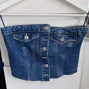 Jeans korsett med knappar - Snygg jeans korsett i blå denim med knappar framtill. Den har en cool, fransig nederkant och dekorativa fickor med knappar. Perfekt för en trendig look!