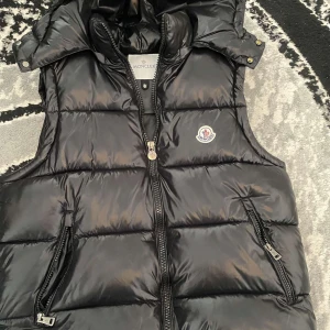 Moncler väst  - Säljer en snygg svart dunväst från Moncler med dragkedja och två fickor framtill. Västen har en quiltad design och Moncler-logga på bröstet. Perfekt för kyligare dagar! Använd 2 gånger bara 