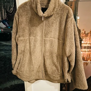 Beige teddyjacka från 157 - Mysig beige teddyjacka från 157 med dragkedja framtill. Perfekt för kyliga dagar med sin fluffiga och varma design. Jackan har långa ärmar och en hög krage för extra värme.