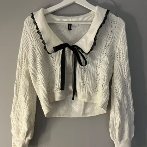 Vit stickad kofta med spetsdetaljer - Charmig vit stickad kofta från H&M med svarta spetsdetaljer och en söt rosett vid kragen. Koftan är croppad och har långa ärmar med ett fint stickat mönster. Säljes då den inte är min stil.