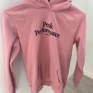 Rosa hoodie från Peak Performance - Säljer en snygg rosa hoodie från Peak Performance med svart text på bröstet. Den har en klassisk känguruficka och en bekväm huva. Perfekt för en avslappnad stil.