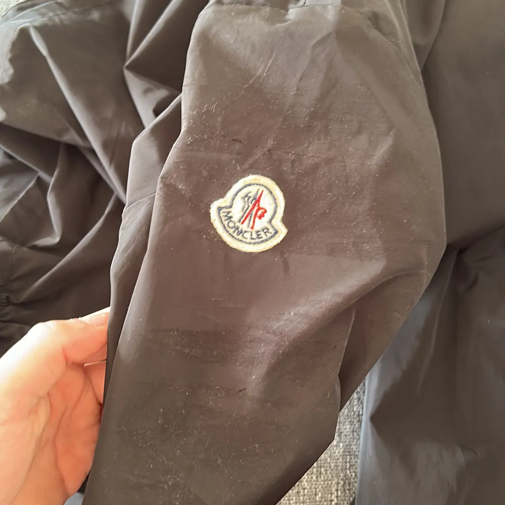 Snygg svart vindjacka från Moncler med dragkedja. Jackan har Moncler-logga på ärmen och detaljer i rött, vitt och blått på huvan. Perfekt för blåsiga dagar. Lite trasig vid moncler märket och på vänster armen( då jag ramlade en gång med jackan). Annars i bra skick. Storlek 2/M men passar 1/S. Takit.