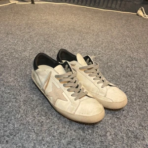 Sneakers från Golden Goose - Säljer ett par stilrena beige sneakers från Golden Goose med en ikonisk stjärndetalj på sidan. Skorna har grå snörning och en svart häl. Sulan är väldigt sliten. Därav priset. Qr kod, låda och dustbag ingår. Fler frågor? Då är det bara att dma mig så har jag svar på det och även fler bilder.