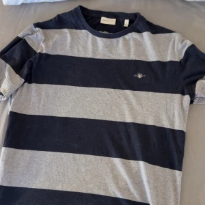 Randig t-shirt från Gant - Säljer en snygg randig t-shirt från Gant i grått och blått Den har korta ärmar och en klassisk rund halsringning. Perfekt för en avslappnad stil. Kvitto finns. Storlek S, Nypris: 550kr.