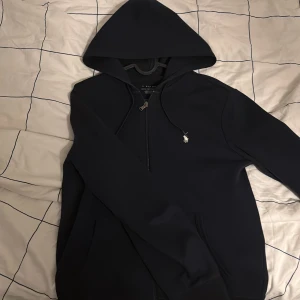 Mörkblå hoodie från Polo - Säljer en stilren mörkblå hoodie från Polo med dragkedja och en liten logotyp på bröstet. Perfekt för en avslappnad look. Den har en klassisk passform och är tillverkad i ett mjukt material.