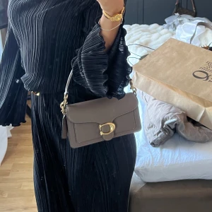 Brun axelväska med gulddetaljer - Snygg brun axelväska med gulddetaljer och ett elegant spänne framtill. Väskan har en justerbar axelrem och en stilren design som passar perfekt för olika tillfällen.