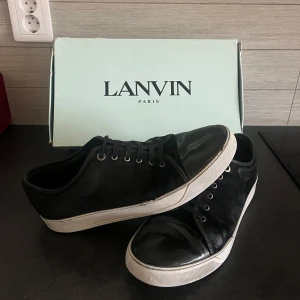 Svarta sneakers från Lanvin - Snygga svarta sneakers från Lanvin med vit sula och snörning. Skorna har en stilren design med en diskret logga på sulan. Perfekta för en avslappnad men elegant look. Allt og medföljs. Storlek 44 passar 44-45