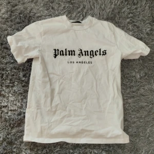Vit t-shirt från Palm Angels - Säljer en vit t-shirt från Palm Angels med tryck i svart text på framsidan. T-shirten har en klassisk rund hals och korta ärmar. Perfekt för en avslappnad stil.