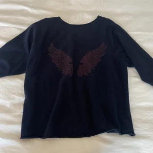 Stickad tröja med vingar - Snygg svart tröja med broderade vingar på ryggen. Tröjan har en offshoulder-design och långa ärmar, vilket ger en avslappnad och trendig look. Perfekt för en stilren outfit🤩🤩