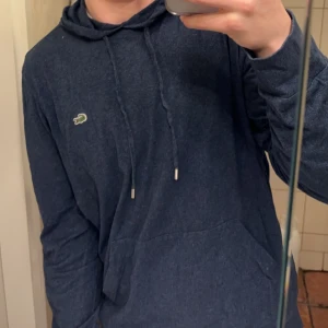 Mörkblå hoodie från Lacoste - Säljer en stilren mörkblå hoodie från Lacoste med den klassiska krokodilloggan på bröstet. Tröjan har en justerbar huva med snören och långa ärmar. Perfekt skick, storlek M men passar även S!