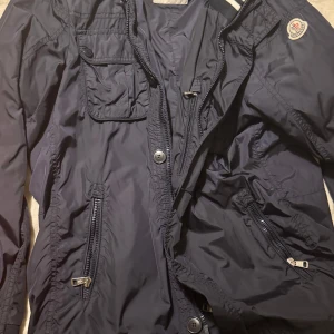 Mörkblå jacka från Moncler - Säljer en snygg mörkblå vintage jacka från Moncler. Jackan har en dragkedja och knappar framtill, samt en bröstficka med knapp. Den har också ribbade detaljer och Moncler-loggan på ärmen. Perfekt för en stilren look! Skicket är 10/10 