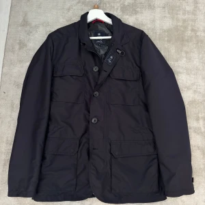 Field Jacket från  Fay - Säljer en stilren svart jacka från Fay med knappar och dragkedja. Jackan har en camouflagemönstrad insida och flera praktiska fickor. Perfekt för höst och vår. Nypris på runt 8000-10 000 kr. 