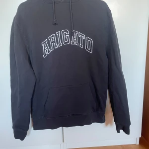 Mörkgrå hoodie från Axel Arigato - Säljer en mörkgrå hoodie från Axel Arigato med texten 'ARIGATO' i vitt broderat. Väldigt bra skick, säljer då den blivit för liten. Storlek S