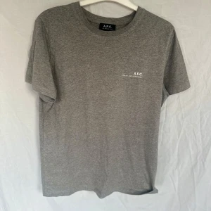 Grå t-shirt från A.P.C. - Säljer en stilren grå t-shirt från A.P.C. med tryck på bröstet. Tillverkad i mjukt material för en bekväm passform. Perfekt för en avslappnad look.