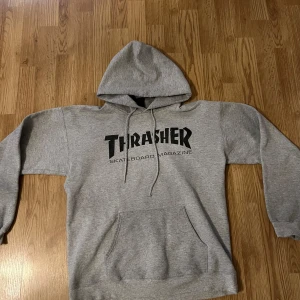 Grå hoodie från Thrasher - Säljer en klassisk grå hoodie från Thrasher med svart tryck på framsidan. Tröjan har en stor ficka framtill och justerbar huva med snören. Perfekt för skate-entusiaster eller som en bekväm vardagströja.