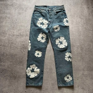 Blå jeans med vita blommor - Unika blå jeans från Levi's med stora vita blommönster över hela byxorna. De har en klassisk femficksdesign och en knappgylf. Perfekta för dig som vill sticka ut med en kreativ stil.
