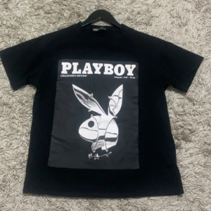 Svart t-shirt med Playboy-tryck - Snygg svart t-shirt med stort Playboy-tryck på framsidan och text på baksidan. Perfekt för en casual look med en touch av stil. T-shirten har korta ärmar och en klassisk passform.