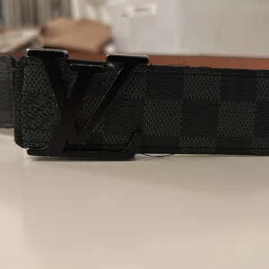 Äkta Rutig bälte från Louis Vuitton - Snyggt bälte från Louis Vuitton med klassiskt rutmönster i svart och grått. Bältet har en stilren LV-spänne i svart och är tillverkat i skinn. Perfekt för att ge en lyxig touch till din outfit.
