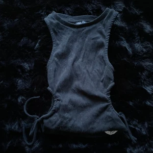 Svart ribbad topp från Zara - Snygg svart ribbad topp från Zara med knytning i sidorna. Perfekt för en stilren look. Toppen är ärmlös och har en tight passform.