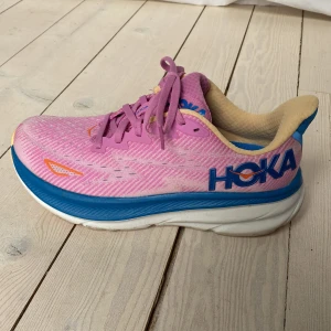 Rosa och blå löparskor från Hoka - Snygga och färgglada löparskor från Hoka i rosa och blått. Modell W Clifton 9. Skorna har en bekväm och dämpad sula, perfekt för långa löprundor. De har snörning och en stilren design med märkets logga på sidan. Blå reflexyta på hälen.  Använda två gånger på promenad men konstaterade att de tyvärr är för små för mig. Storlek 38 men känns mer som 37. Pris t ex på Stadium 1899 kr. 