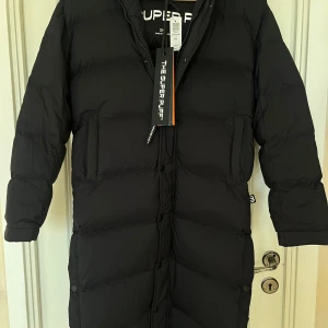 Svart lång dunjacka från Aritzia - Helt ny med prislapp! Säljer en svart lång dunjacka från Aritzia. Jackan har en pufferdesign med långa ärmar och en praktisk huva. Perfekt för kalla vinterdagar med sin varma och stilrena look. Den har en dragkedja framtill och är i en klassisk svart färg.