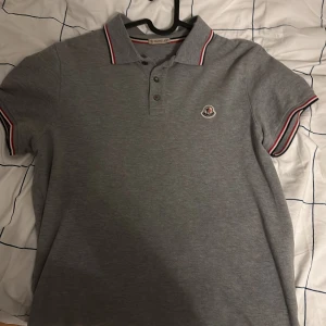 Grå pikétröja från Moncler - Snygg grå pikétröja från Moncler med klassisk krage och knappar framtill. Tröjan har korta ärmar med röd och vit randig detalj vid ärmslut och krage. Perfekt för en stilren look.