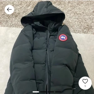 Svart dunjacka med huva.  - Säljer en svart dunjacka med huva och dragkedja. Jackan har en rund logotyp på bröstet och är perfekt för kalla dagar. Den är stilren och funktionell.