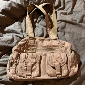 Beige/Brun axelväska från Gabaara - Säljer en snygg beige/brun axelväska från Gabaara. Väskan har två framfickor med knappar och dragkedja upptill. Insidan är mönstrad med ett stilrent tryck. Man kan ha hur mycket som helst i väskan, den är så rimlig. Den har hela sex fack på utsidan och tre på insidag. Otroligt bra rymlig väska!