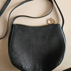 Svart axelväska med dragkedja - Snygg svart axelväska i syntetmaterial med en praktisk dragkedja upptill. Väskan har en elegant och enkel design med en smal axelrem och en liten kedjedetalj. Perfekt för att bära med sig det viktigaste på språng.