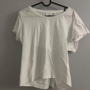 Vit t-shirt med knutdetalj - Säljer en vit t-shirt med en unik knutdetalj på ryggen som skapar en öppen rygg. T-shirten har korta ärmar och är tillverkad i ett mjukt bomullsmaterial. Perfekt för en avslappnad stil.