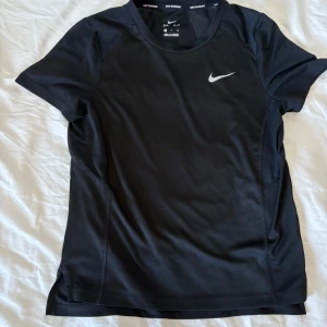 Svart tränings-t-shirt från Nike - Snygg svart tränings-t-shirt från Nike med korta ärmar och en diskret logga på bröstet. Perfekt för gymmet eller löprundan. Tillverkad i ett lätt och andningsbart material för maximal komfort under träning. Sista bilden är mer för att visa hur den ser ut på.