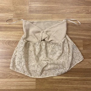 Beige axelbandslös topp med spetsdetaljer - Charmig beige axelbandslös topp med en stickad överdel och knytdetalj framtill. Nederdelen har vacker spets med blommönster. Perfekt för varma dagar och en stilren look.