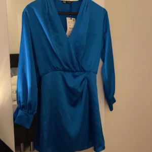 Blå omlottklänning från Zara - Blå omlottklänning från Zara med långa ärmar och v-ringning. Klänningen har en mjuk och glansig.