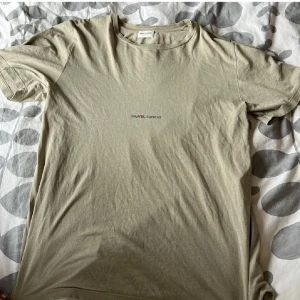 Beige t-shirt från Saint Laurent - Säljer en stilren beige t-shirt från Saint Laurent med korta ärmar. T-shirten har en enkel design med märkesloggan tryckt på bröstet. Perfekt för en avslappnad look.