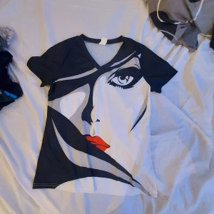 Svart och vit t-shirt med ansiktsmotiv - Snygg t-shirt med ett stort ansiktsmotiv i svart och vitt, med röda läppar som detalj. Tillverkad i polyester och spandex för en bekväm passform. Perfekt för dig som vill sticka ut med en unik design.