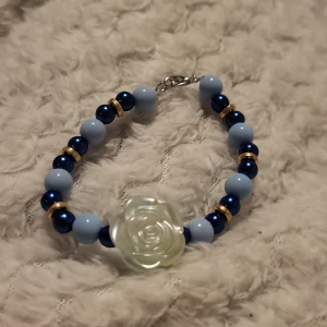Armband med blå pärlor och rosdetalj - Charmigt armband med ljusblå och mörkblå pärlor, dekorerat med guldfärgade detaljer. Mittpunkten är en vacker vit ros. Perfekt för att ge en färgklick till din outfit.