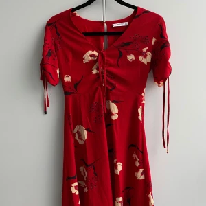 Röd blommig klänning från Mango - Säljer en superfin röd klänning från Mango med blommigt mönster i beige och svart. Klänningen har korta ärmar med snörning och en v-ringad hals. Perfekt för sommarens fester eller en dag på stan!