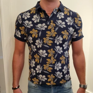 Pikétröja med blommigt mönster - Pikétröja av 100% bomull, perfekt för sommriga dagar. Den har normal passform, med knappar i kragen och klassisk ribbstickning i ärmar och krage. Har använts ett fåtal gånger. Bra skick. 