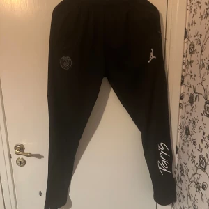Svarta mjukisbyxor med PSG-logga - Säljer ett par svarta mjukisbyxor med Paris Saint-Germain-logga och Jumpman-märke. Byxorna har en avslappnad passform och är perfekta för både träning och chill. De har elastisk midja och är gjorda i ett mjukt material för extra komfort. På ena benet står det 'Paris' i vit text.