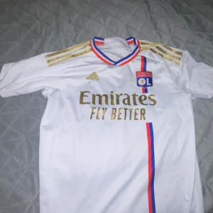 Vit fotbollströja från Adidas - Säljer en snygg vit fotbollströja från Adidas med guldiga detaljer och ränder på axlarna. Tröjan har korta ärmar och en cool design med klubbmärke och sponsorlogga på framsidan. Perfekt för fotbollsfans som vill ha en stilren look!