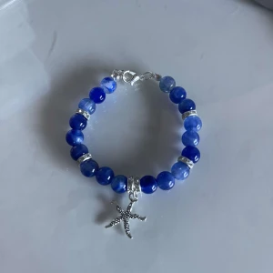 Blått armband med sjöstjärneberlock - Snyggt armband med blå pärlor och en söt sjöstjärneberlock i silver. Perfekt för att ge en marin touch till din outfit. Armbandet har en enkel låsning och passar de flesta handleder.